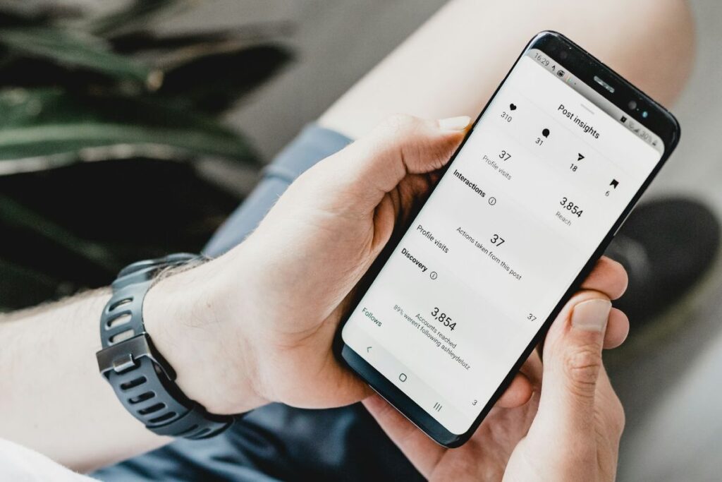 Person's hands holding a smartphone showing social media post insights and analytics, with a black fitness watch on the wrist.