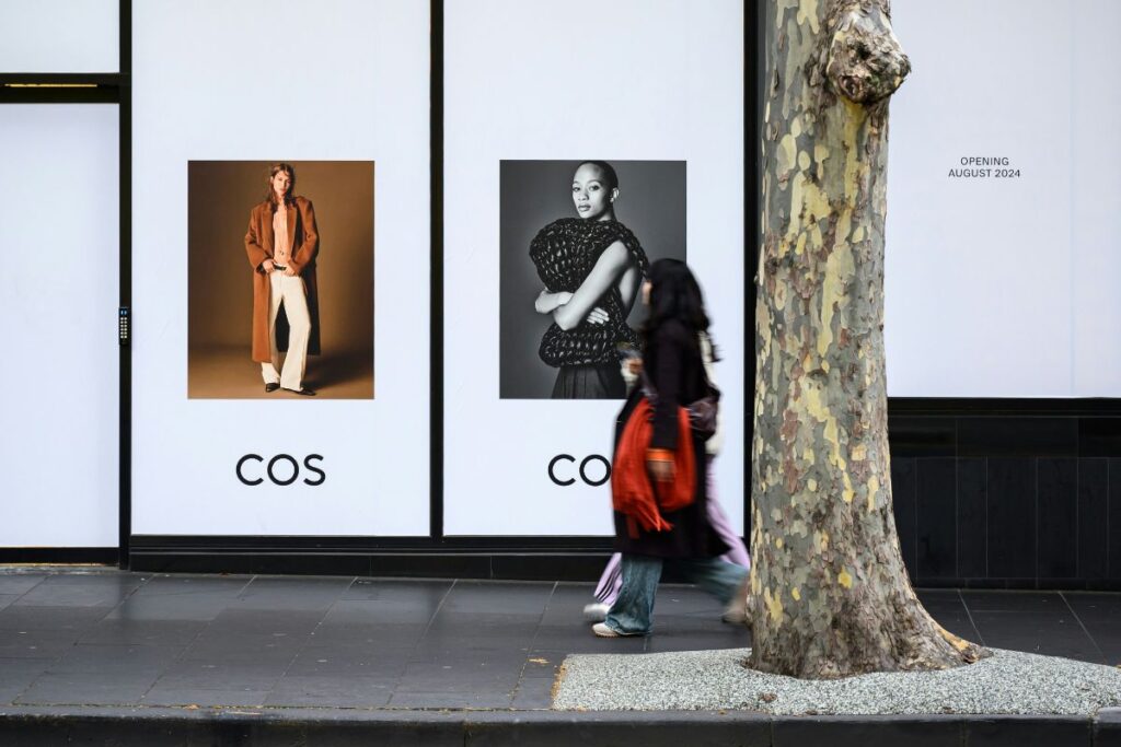 Storefront hoarding with two COS fashion posters, a blurred pedestrian carrying an orange bag walking past and a plane tree trunk.