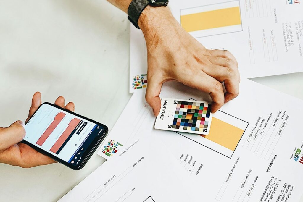 Hand holding a Pantone color swatch over printed layouts while another hand holds a smartphone displaying color samples.